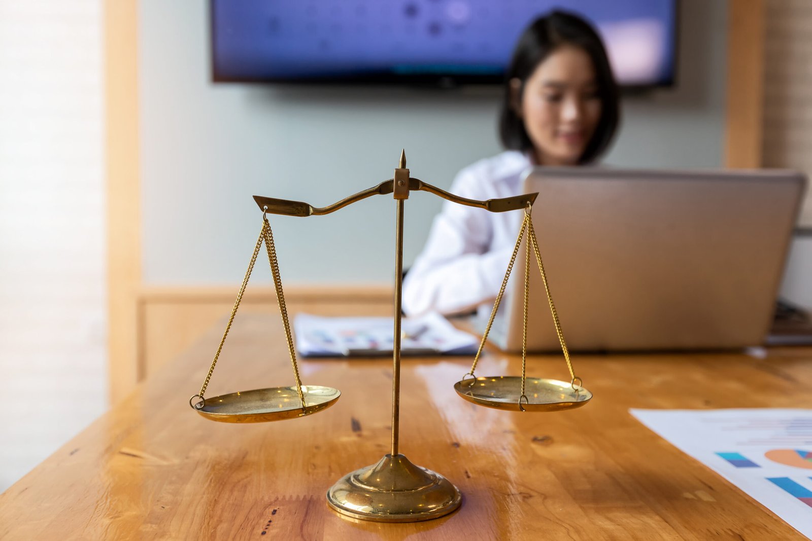 Golden scales of justice on a desk with a person working on a laptop in the background.