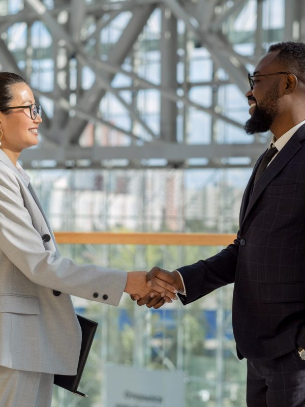 Happy young intercultural colleagues shaking hands while standing in front of camera
